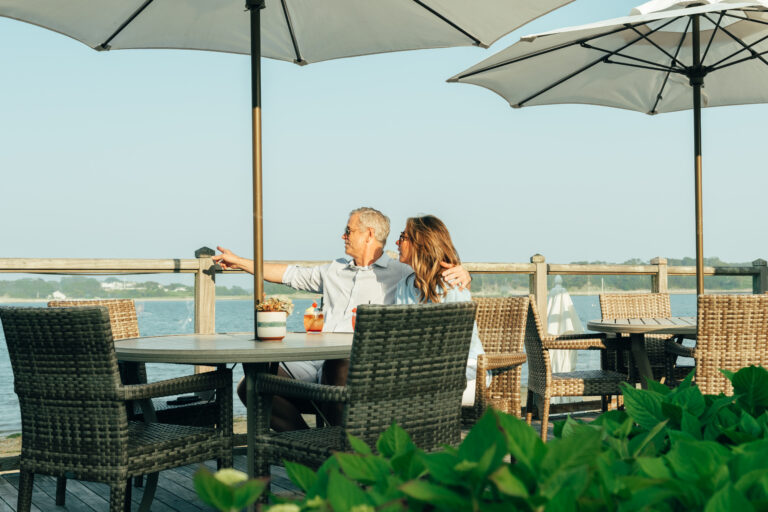 An older man and woman sit together at an outdoor table by the water under umbrellas at a restaurant Cape Cod, with the man pointing toward the distance.