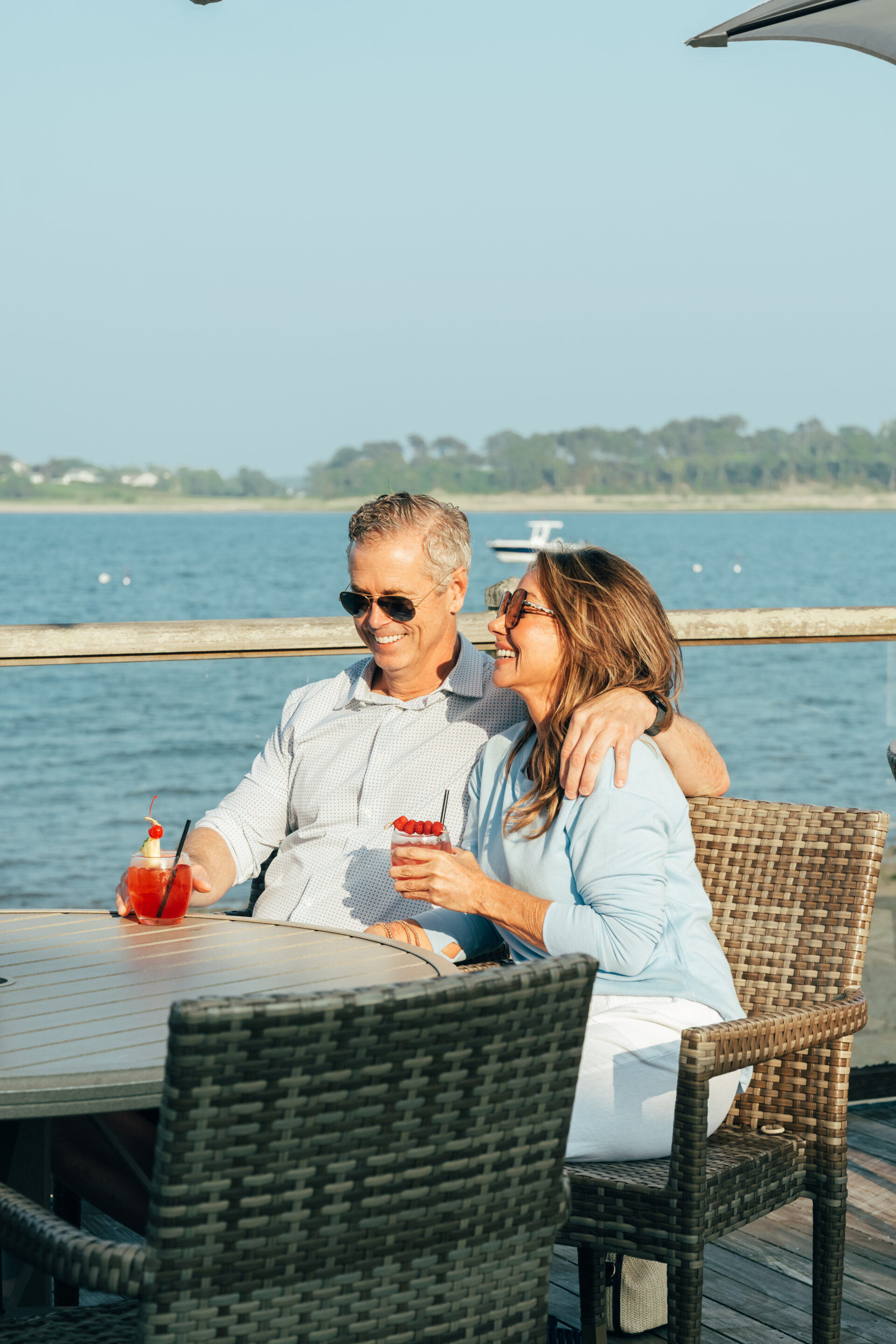 A man and woman sit together at an outdoor table by the water, smiling and holding drinks with garnishes on a sunny day at Wequassett Resort and Golf Club.