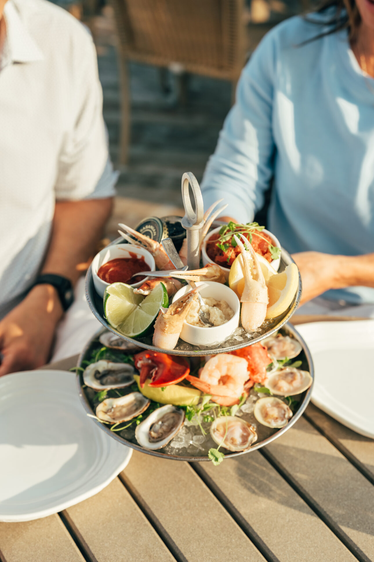 Two people sit at a table with a two-tiered seafood platter featuring oysters, shrimp, crab claws, lemon wedges, and dipping sauces at a charming Cape Cod hotel.