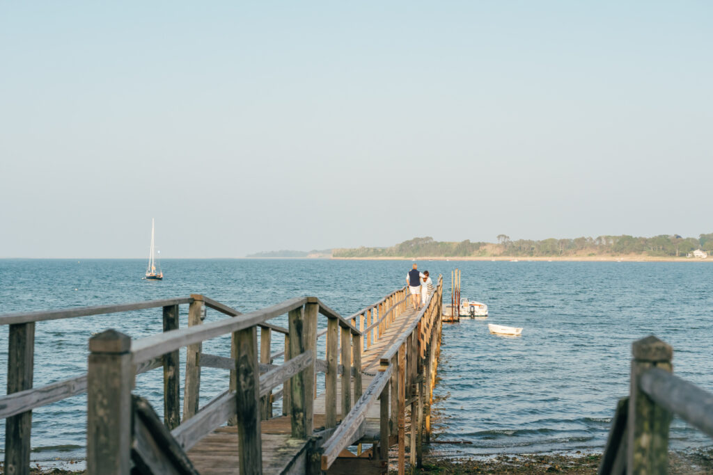 A wooden pier extends into the ocean at Wequassett Resort and Golf Club with a couple walking at the end; small boats float nearby and a sailboat drifts in the distance under a clear sky.