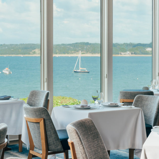 A restaurant dining area with tables and chairs near large windows overlooking a calm body of water with sailboats and a distant shoreline.