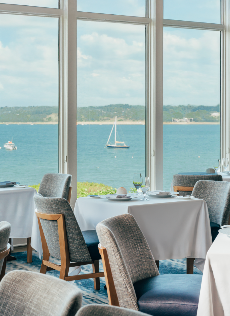 A restaurant dining area with tables and chairs near large windows overlooking a calm body of water with sailboats and a distant shoreline.