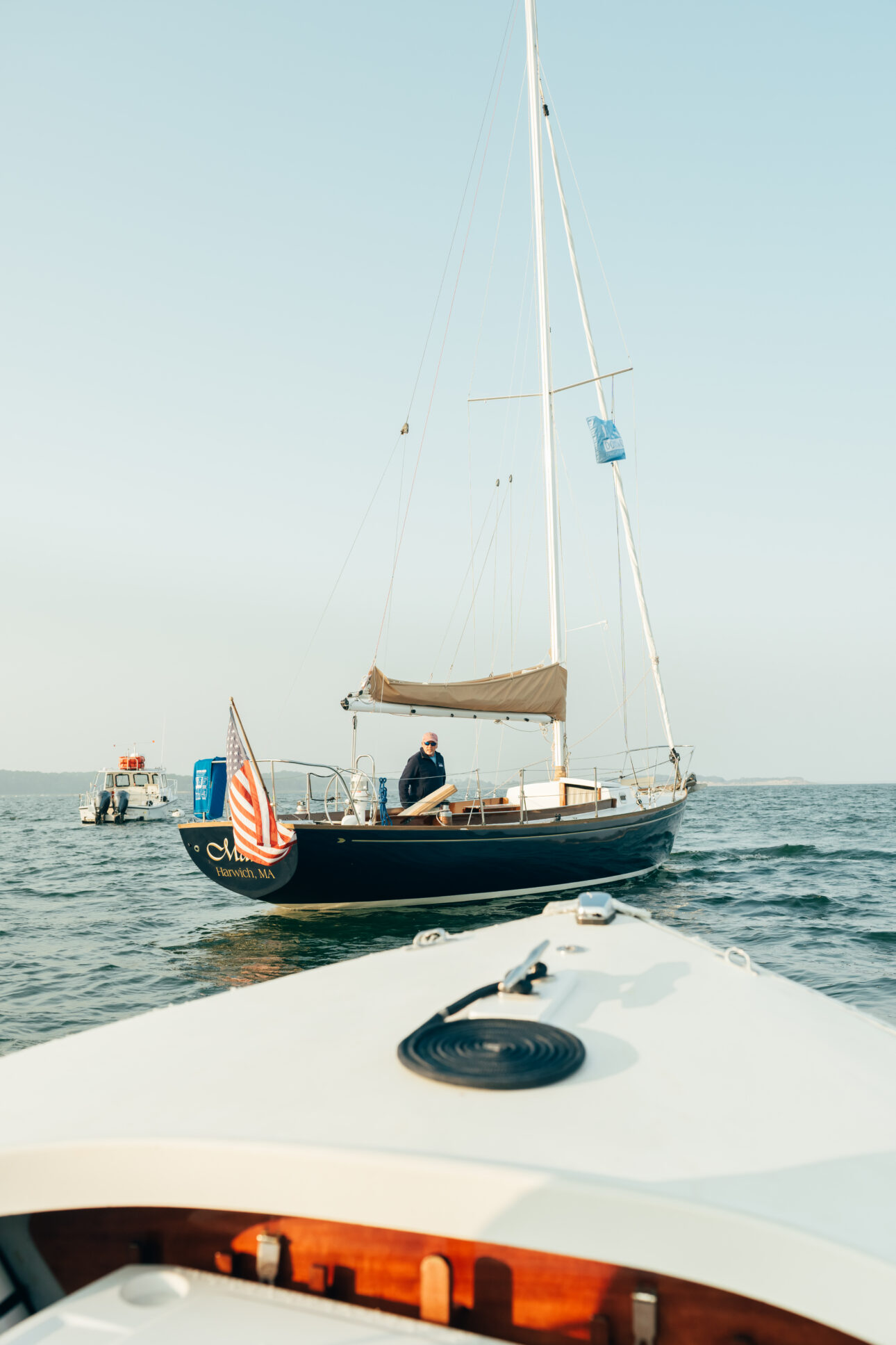 A sailboat with a person onboard and an American flag at the stern is seen from another boat on calm water under a clear sky near Wequassett Resort and Golf Club, one of the top Cape Cod resorts.
