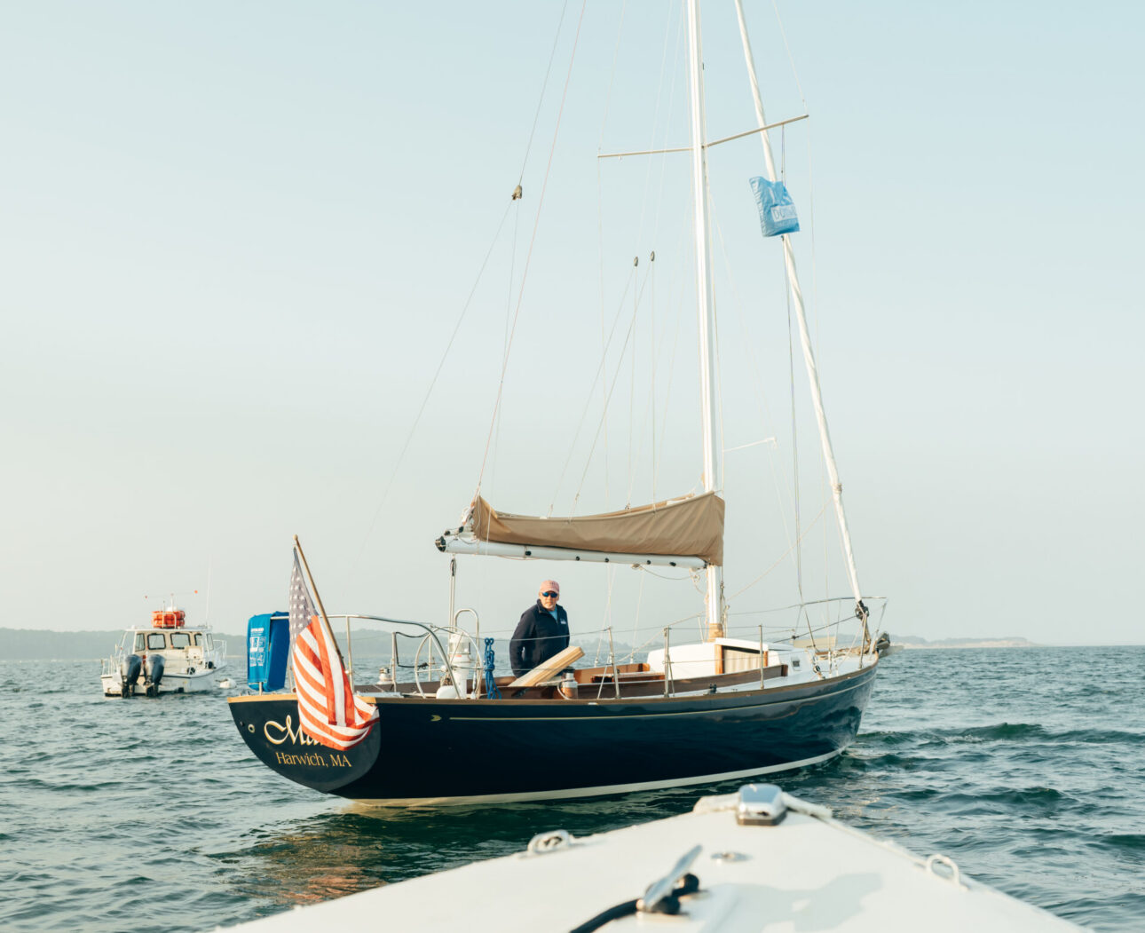 A sailboat with a person onboard and an American flag at the stern is seen from another boat on calm water under a clear sky near Wequassett Resort and Golf Club, one of the top Cape Cod resorts.