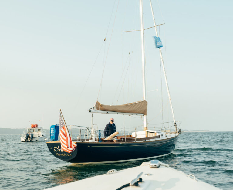 A sailboat with a person onboard and an American flag at the stern is seen from another boat on calm water under a clear sky near Wequassett Resort and Golf Club, one of the top Cape Cod resorts.