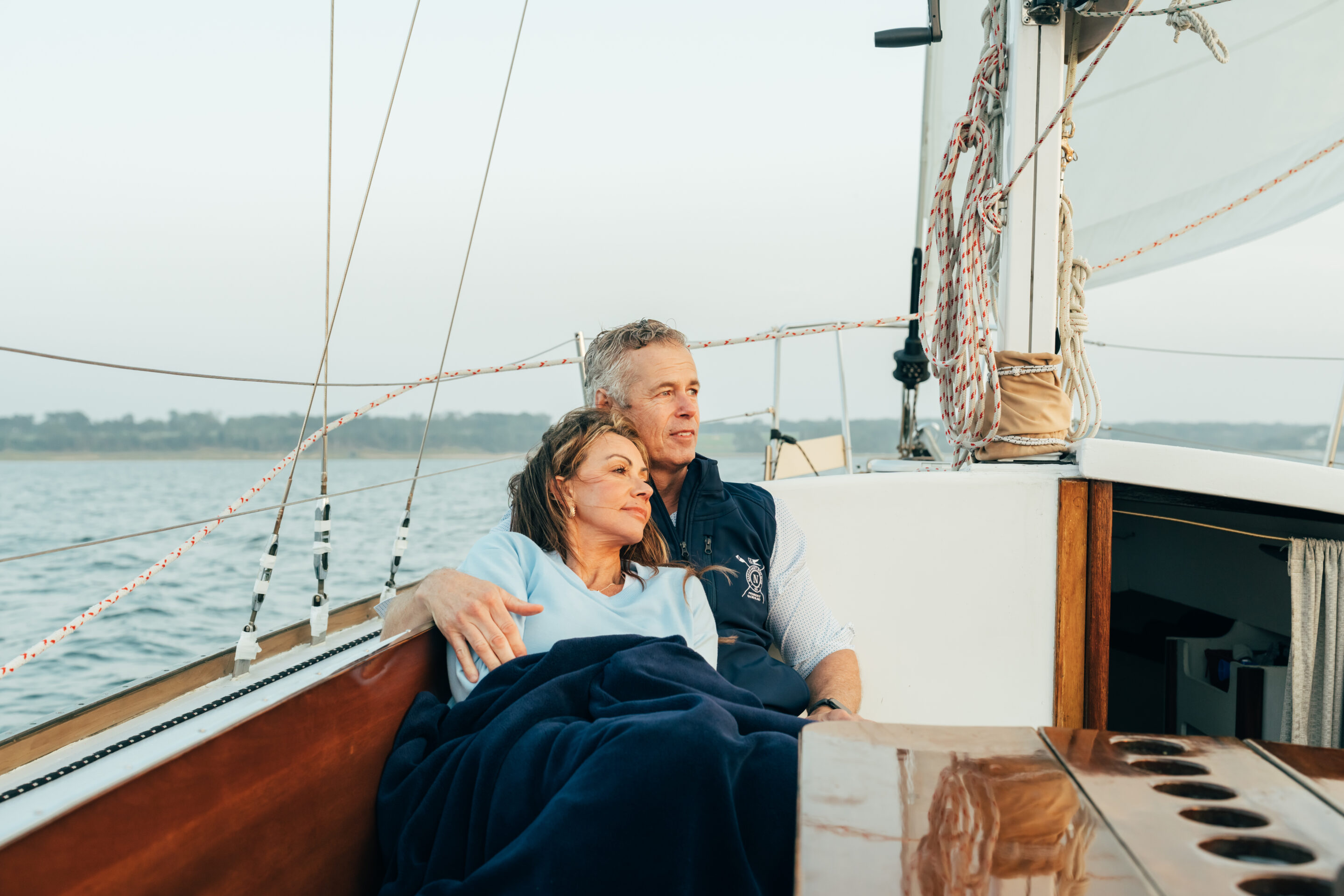 A man and woman sit closely together, wrapped in a dark blanket, relaxing on the deck of a sailboat with water and the shoreline near the Wequassett resort in the background.