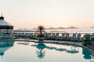 A poolside area with lounge chairs and umbrellas at Wequassett Resort overlooks a calm body of water at sunrise or sunset, with a gazebo and palm plants nearby—perfect for a relaxing getaway from NYC.