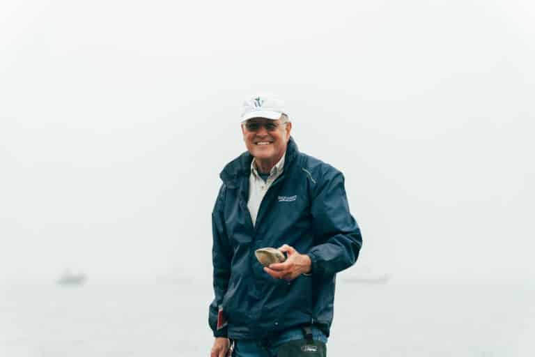 Captain John, an older man in a blue jacket and white cap, stands outdoors near water, smiling and holding a clam during one of his clamming tours with a foggy background.