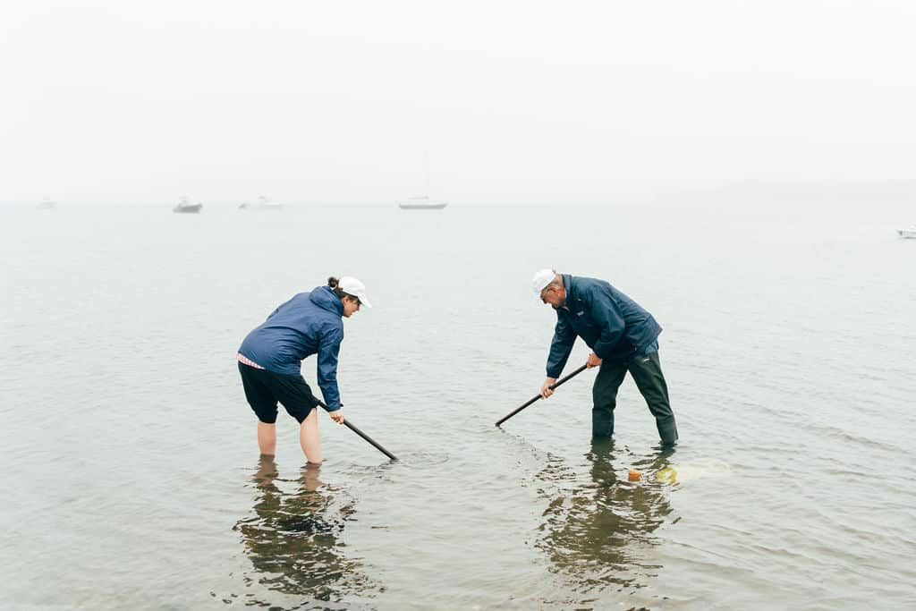 Two people wearing jackets and hats stand in shallow water, clamming with sticks in the mud on a foggy day; boats are visible in the background, evoking scenes reminiscent of Captain John's coastal adventures.
