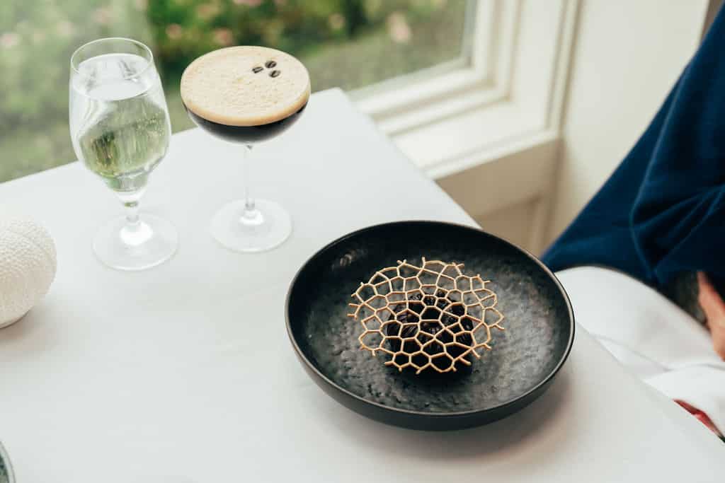 A glass of water, an espresso martini, and a black plate with a decorative honeycomb-patterned garnish on a white tablecloth near a window set the scene for fine dining at this Cape Cod restaurant.