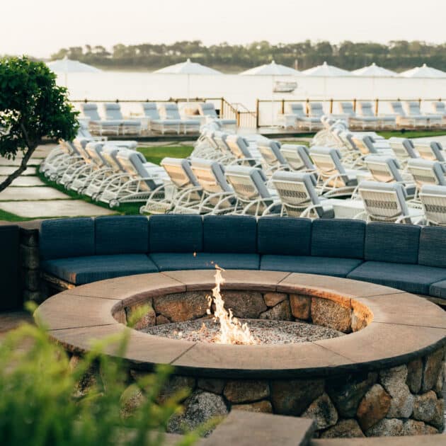 A stone fire pit with a small flame is surrounded by cushioned seating, with rows of empty lounge chairs and tents in the background near a body of water.