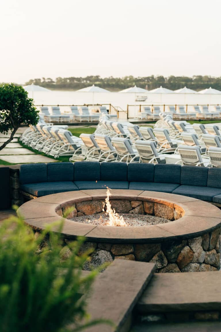 A stone fire pit with a small flame is surrounded by cushioned seating, with rows of empty lounge chairs and tents in the background near a body of water.