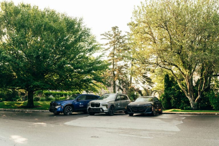 Three parked BMW vehicles—blue, silver, and black—are lined up side by side on a residential street, surrounded by green trees and landscaping. Discover these models and current special offers available now.
