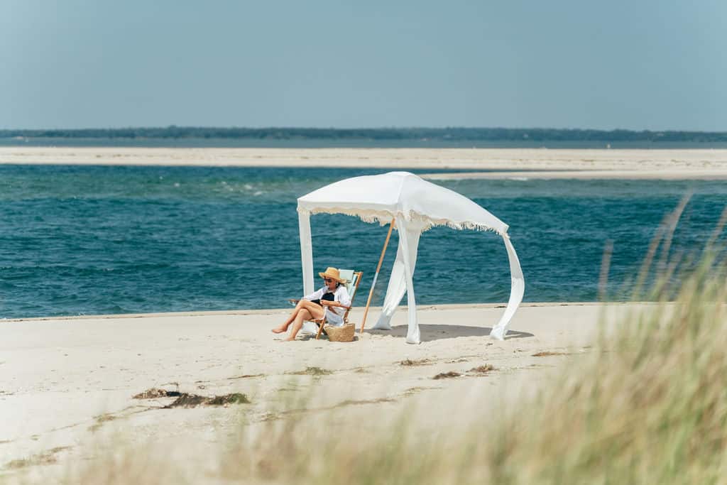 A person embarks on a beach journey, sitting under a white canopy on Outer Beach’s sandy shore, wearing a sunhat and holding a book. Grasses sway gently in the foreground.