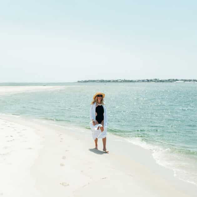 A person wearing a sunhat, sunglasses, and light clothing stands on a sandy beach near the edge of the water on a bright, clear day.