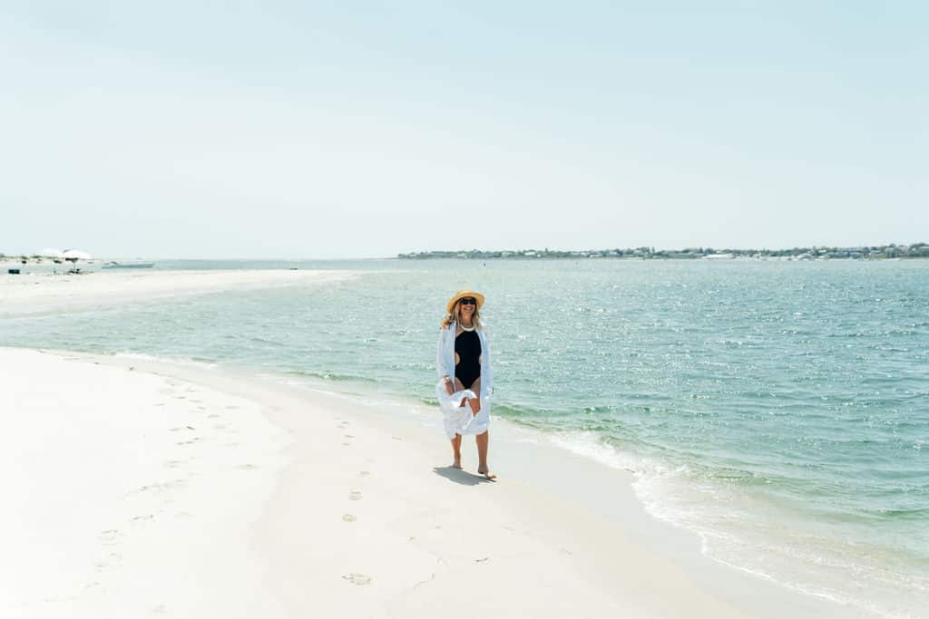 A person wearing a sunhat, sunglasses, and light clothing stands on a sandy beach near the edge of the water on a bright, clear day.
