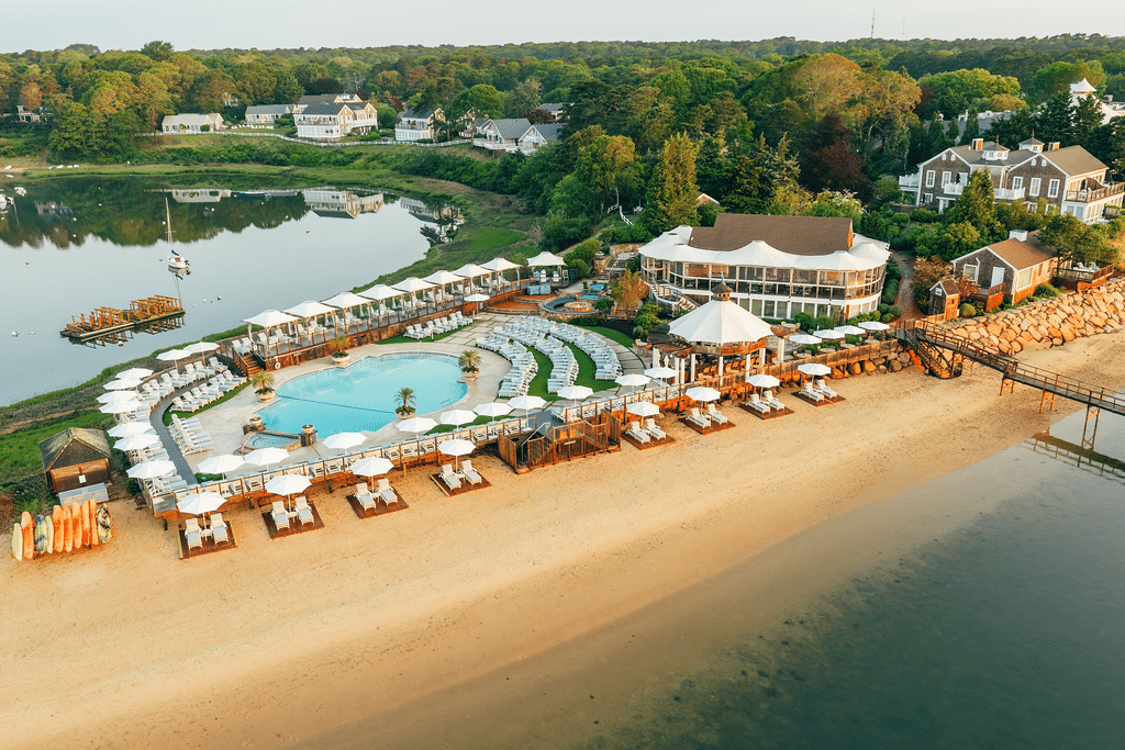Aerial view of Pleasant Bay Explorers waterfront resort with a pool, lounge chairs, umbrellas, and beachfront cabanas, surrounded by greenery and residential houses near the calm waters of Pleasant Bay.