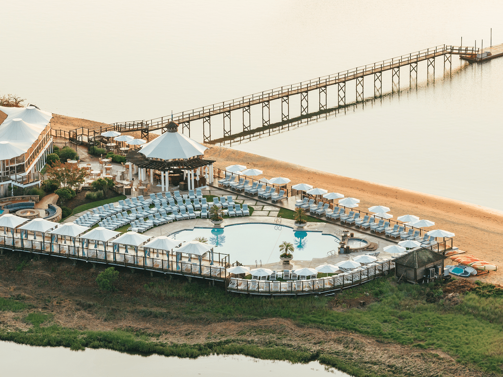 Aerial view of a resort pool area with lounge chairs and umbrellas near a sandy beach and a long wooden pier extending into calm water, radiating an atmosphere of Everlasting Elegance.