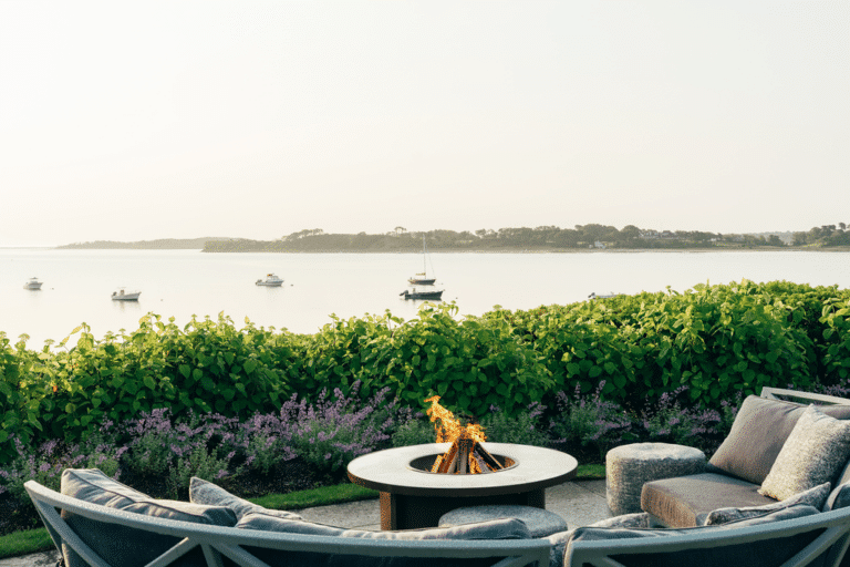 Outdoor seating area with a lit fire pit overlooks calm water with several anchored boats and lush greenery in the background.