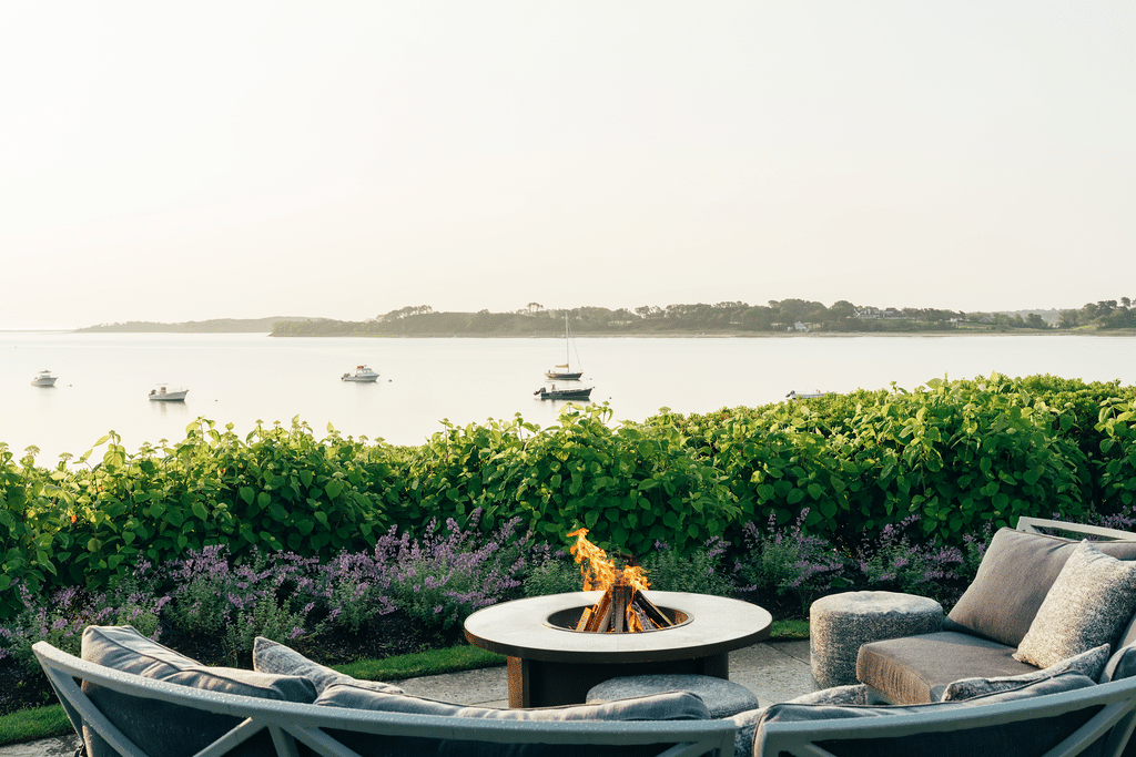 Outdoor seating area with a lit fire pit overlooks calm water with several anchored boats and lush greenery in the background.