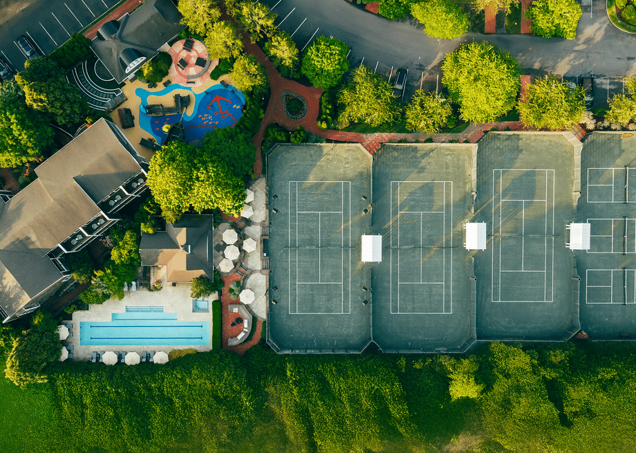 Aerial view of a sports complex featuring tennis courts for racquet sports, a swimming pool with poolside umbrellas, a playground, buildings, and lush surrounding greenery.
