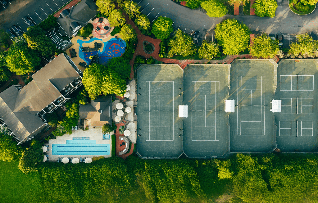 Aerial view of a sports complex featuring tennis courts for racquet sports, a swimming pool with poolside umbrellas, a playground, buildings, and lush surrounding greenery.