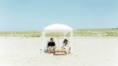 Two people sit under a white canopy on the Outer Beach, surrounded by sandy stretches and grassy dunes, wearing sunglasses and relaxing in wooden chairs—enjoying a peaceful moment on their coastal journey.