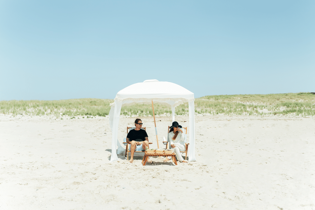 Two people sit under a white canopy on the Outer Beach, surrounded by sandy stretches and grassy dunes, wearing sunglasses and relaxing in wooden chairs—enjoying a peaceful moment on their coastal journey.