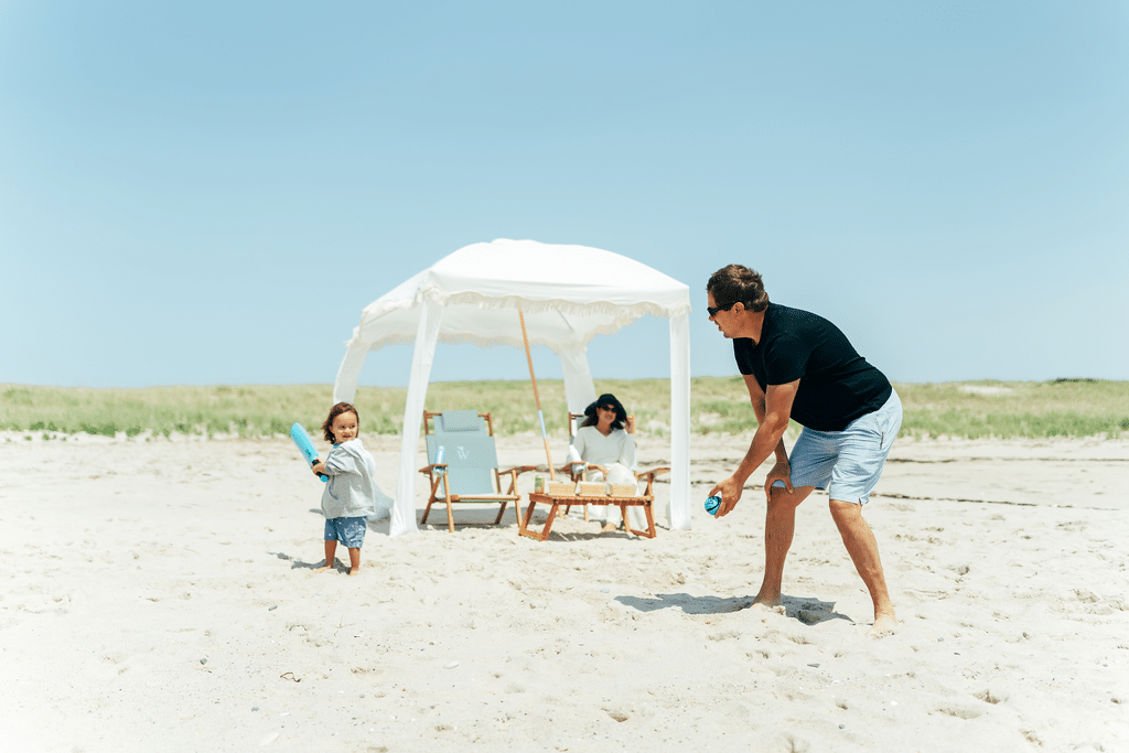A man and a child enjoy family fun with plastic bats on a sandy beach, while a woman sits under a white canopy in the background.