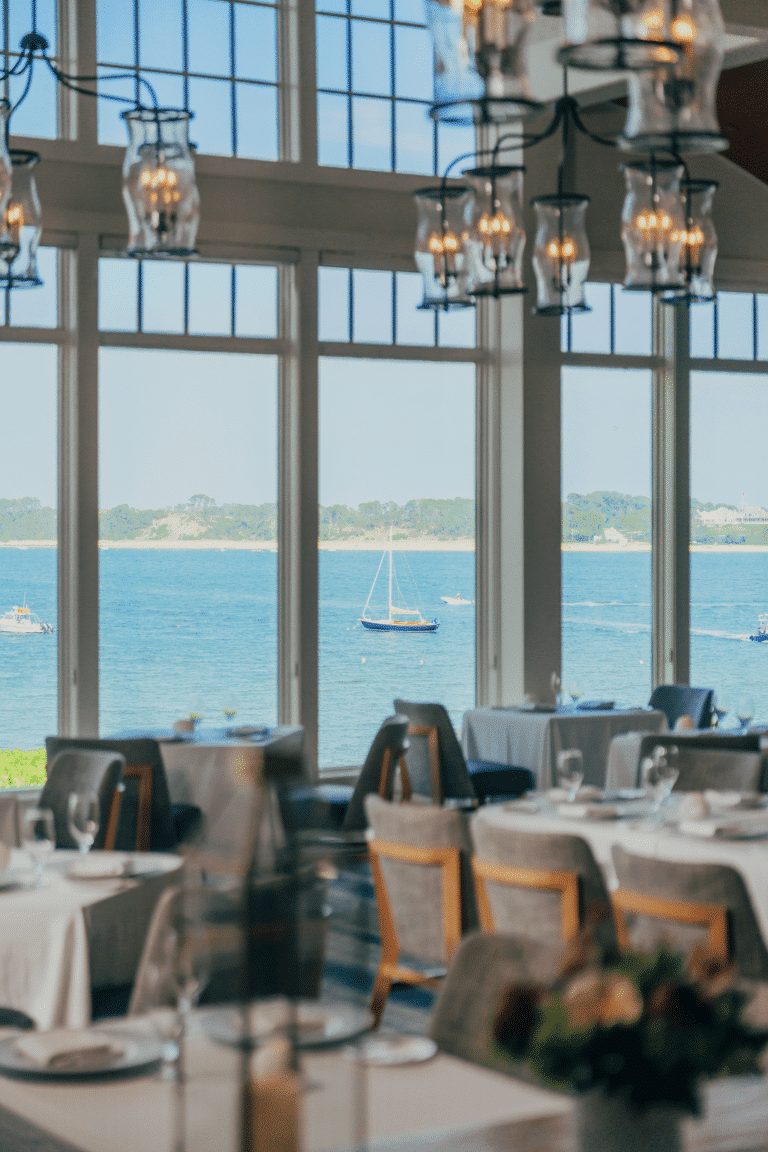 Tables set for dining in a restaurant with large windows overlooking a calm bay with sailboats and greenery in the background.