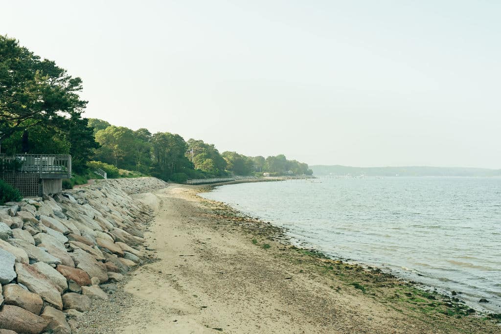 A rocky shoreline with a sandy beach and calm water, bordered by trees and a wooden fence on the left side.