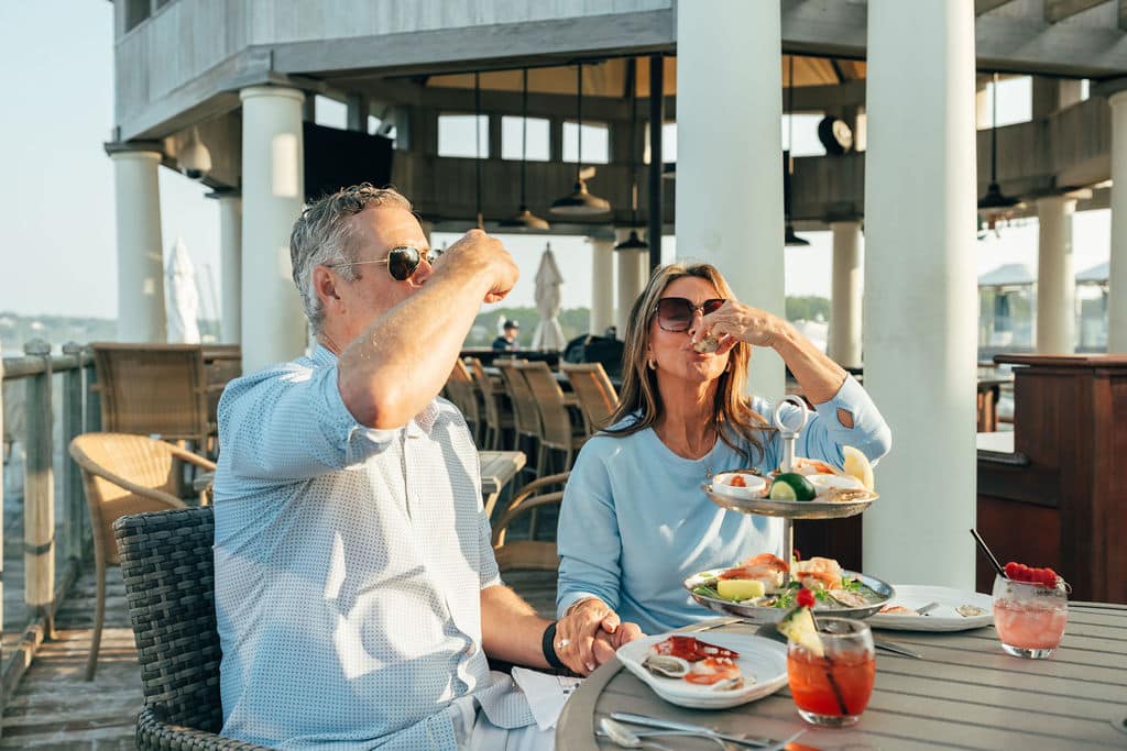 A man and woman sit at an outdoor restaurant table, eating seafood and drinking cocktails in the sunlight.