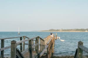 A wooden pier extends over the water with two people standing at the end; a sailboat and small boats in the distance capture classic On the Water Experiences on a calm, sunny day.