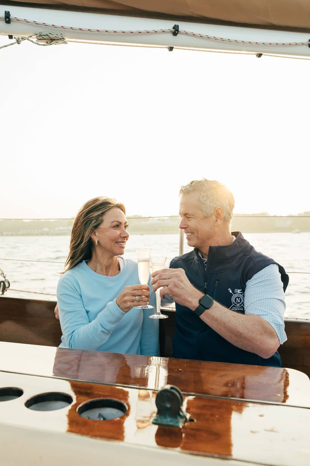 A man and woman sit on a boat, smiling and clinking champagne glasses, enjoying luxurious On the Water Experiences with sparkling sunlight in the background.