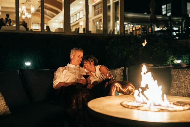 A couple sits close together on an outdoor sofa at The Verandah, drinking wine by a fire pit at night, with a building lit up in the background.