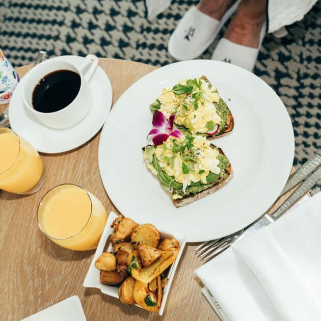 A breakfast table with avocado toast, roasted potatoes, two glasses of orange juice, a cup of coffee, a children's cup, and pancakes. Person's feet in slippers are visible in the background.