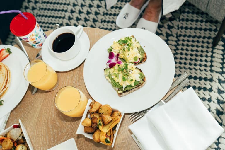 A breakfast table with avocado toast, roasted potatoes, two glasses of orange juice, a cup of coffee, a children's cup, and pancakes. Person's feet in slippers are visible in the background.
