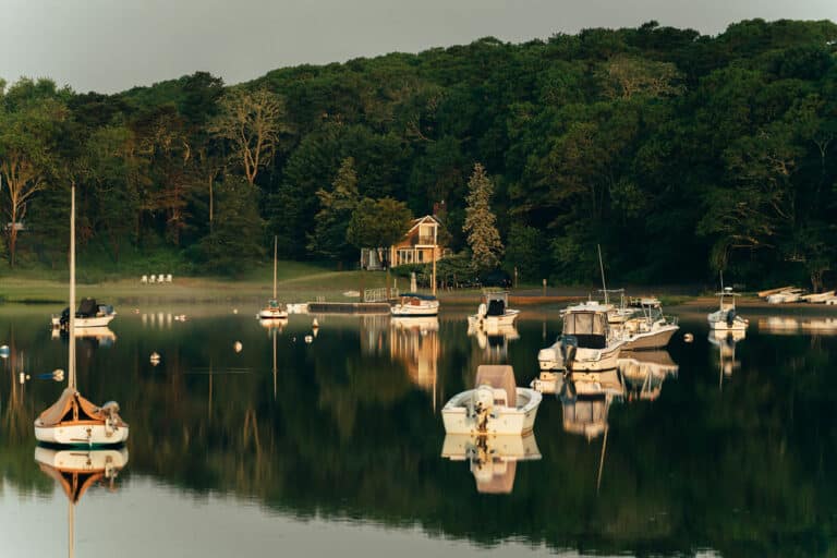 Several boats are anchored on calm water reflecting a forested shoreline with a single house partially visible among dense green trees near the renowned Wequassett Resort and Golf Club.