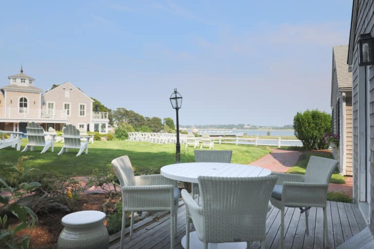 Outdoor patio with a round white table and chairs, overlooking a grassy lawn with Adirondack chairs at a Cape Cod hotel, a building, and water in the background under a clear sky.