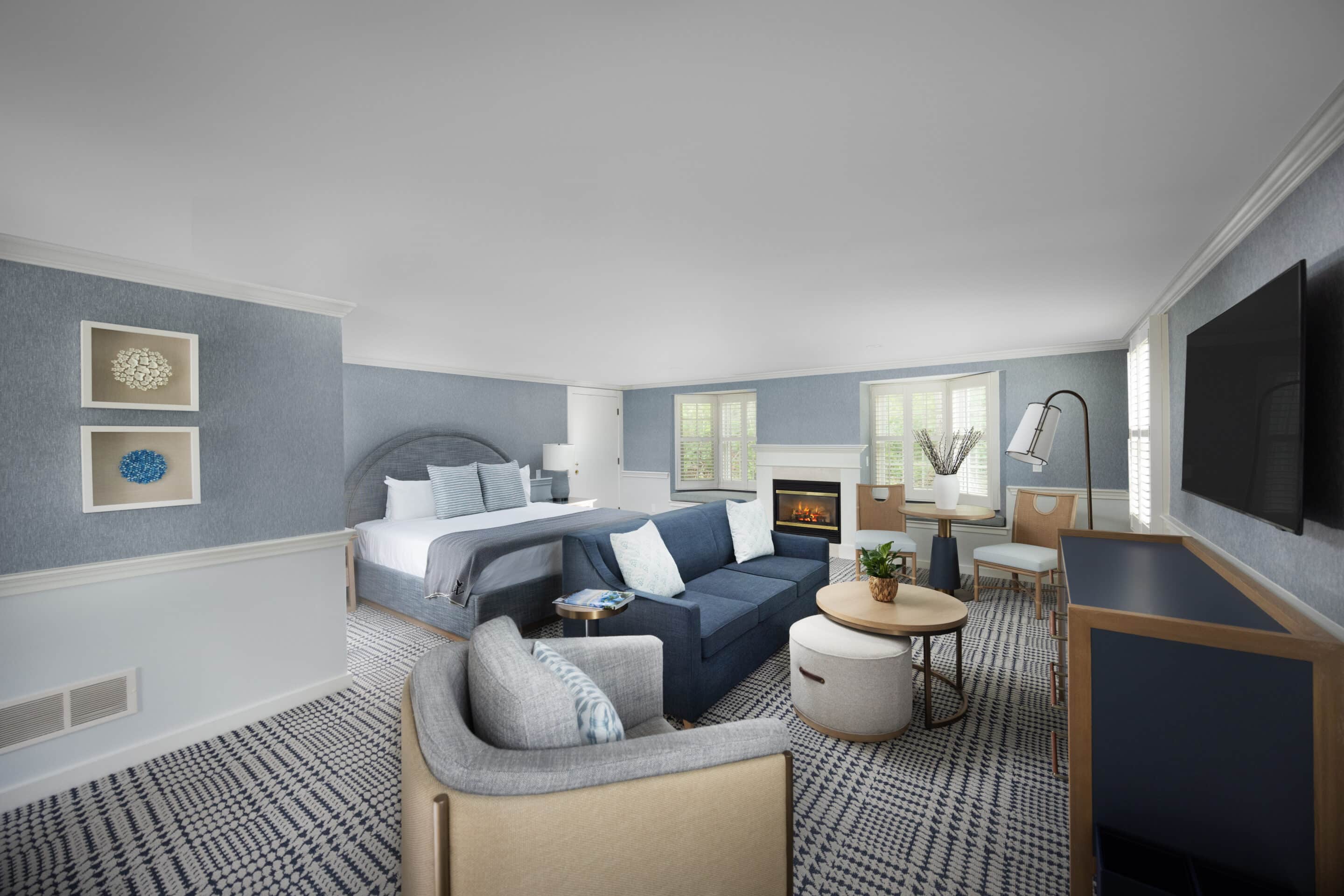 Spacious hotel room with a king bed, blue sofa, armchair, round table, flat-screen TV, fireplace, and large windows in soothing neutral and blue tones—perfect for relaxing after a day at the Cape Cod golf course.