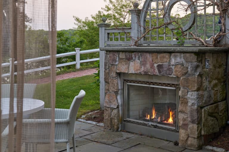 Outdoor stone fireplace with a fire burning next to a patio area with a round white table and chairs, surrounded by greenery and a white fence—perfect for gatherings at 28 Atlantic, an elegant Cape Cod wedding venue.