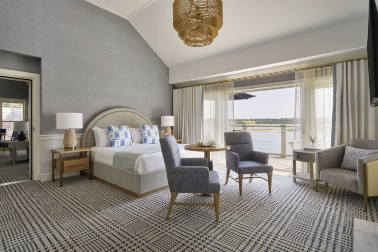 A modern Cape Cod hotel room with a large bed, two blue chairs, a small table, armchair, lamps, and sliding doors opening to a balcony with water views at Wequassett Resort and Golf Club.