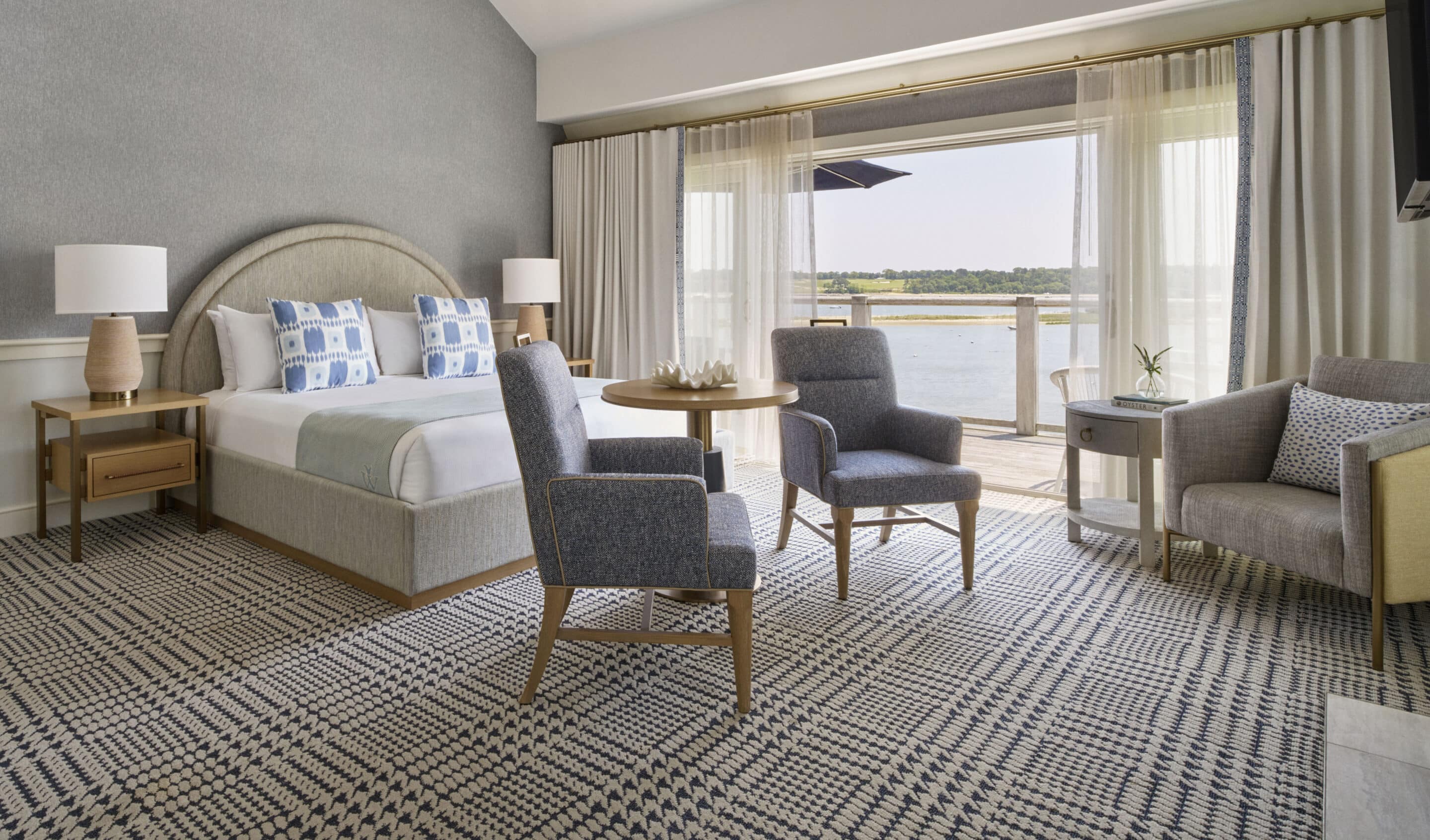 A modern Cape Cod hotel room with a large bed, two blue chairs, a small table, armchair, lamps, and sliding doors opening to a balcony with water views at Wequassett Resort and Golf Club.
