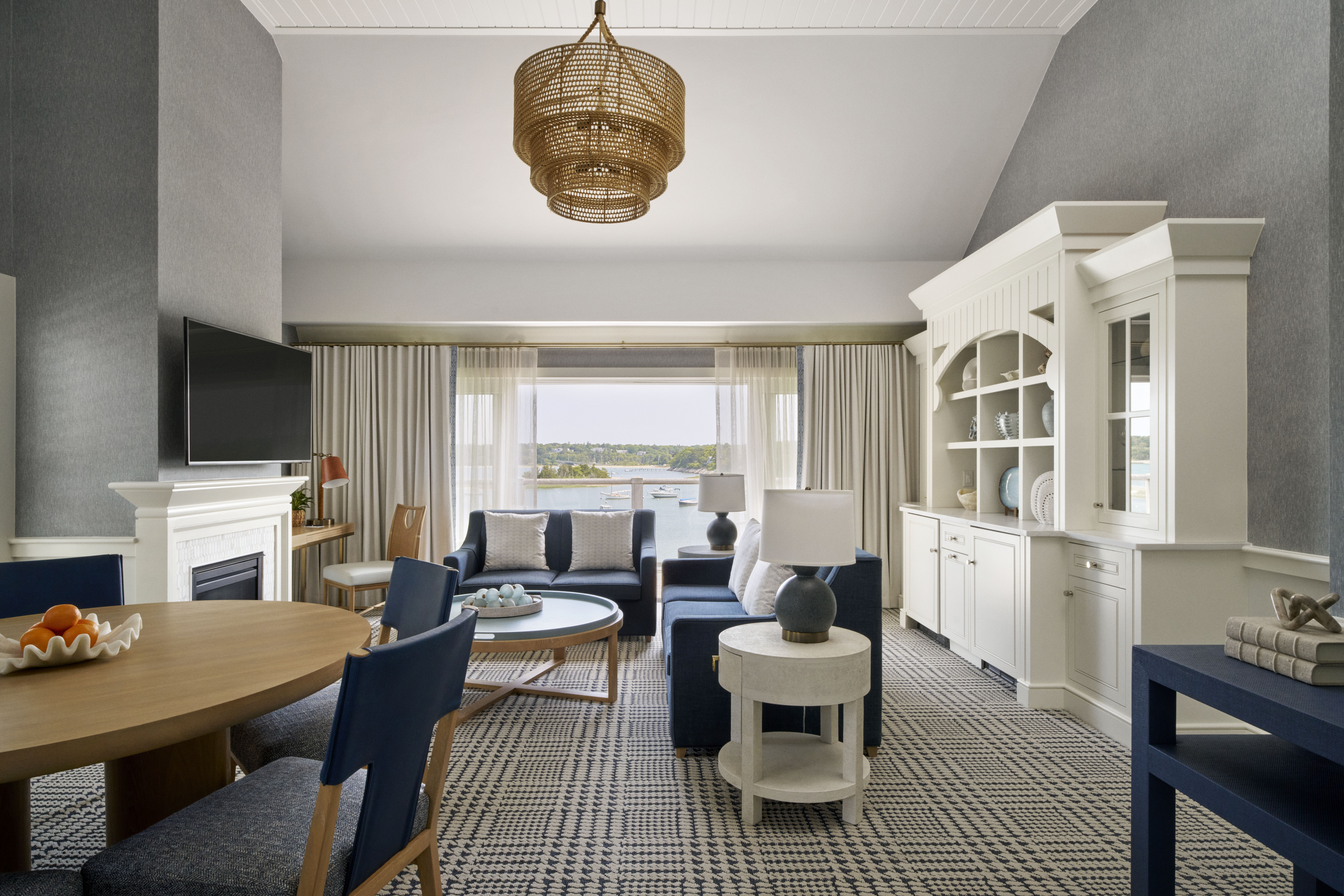 A modern living and dining area with blue and white furniture, a round dining table, wall-mounted TV, large windows with curtains, and a view of greenery—perfect for relaxing after a day on the Cape Cod golf course.