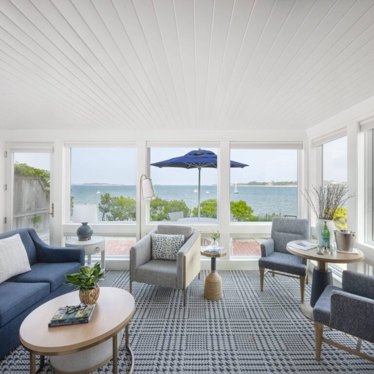 Bright living room with modern furniture, large windows overlooking a waterfront view, and a patio with a blue umbrella outside at the scenic Wequassett Resort and Golf Club—an ideal Cape Cod wedding venue.