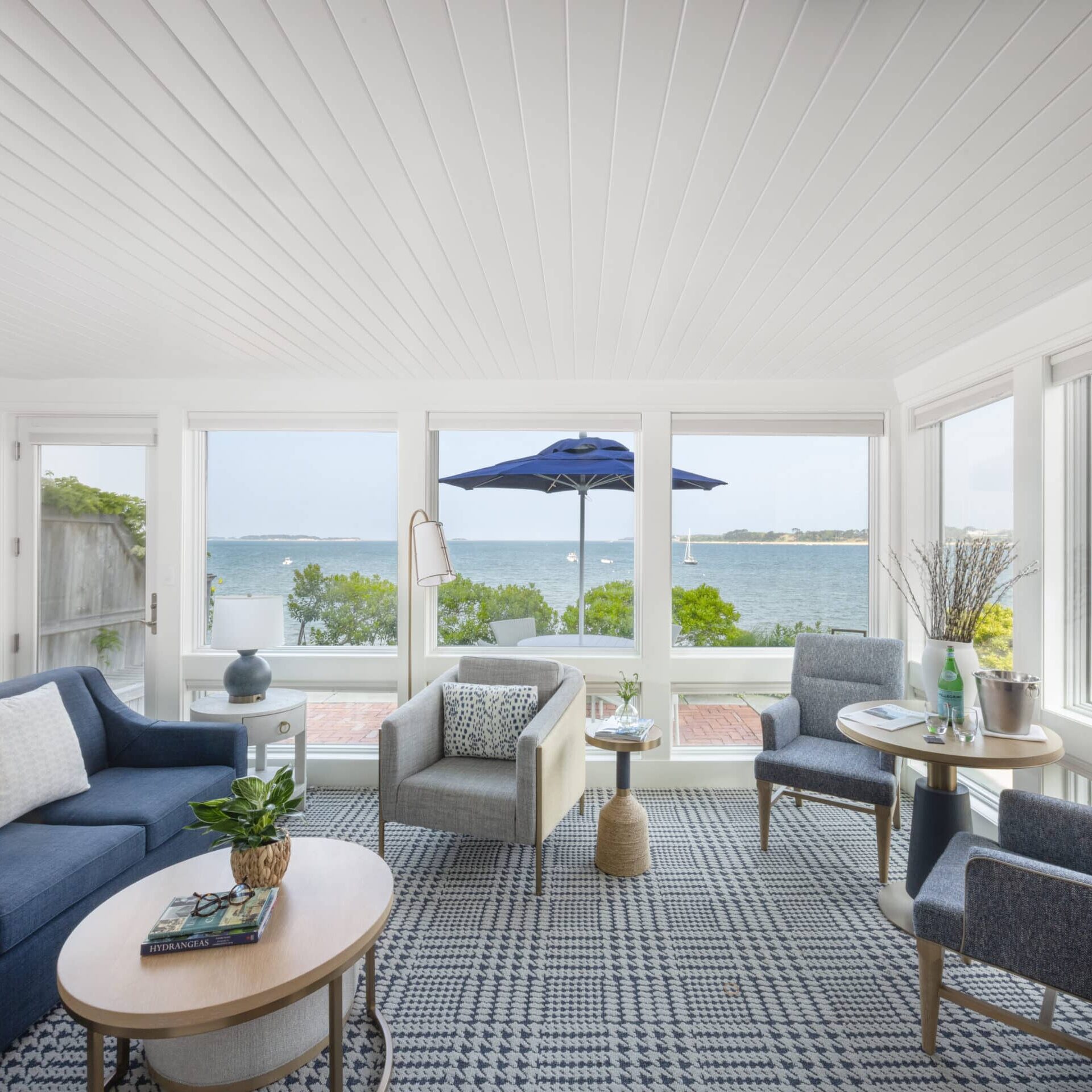 Bright living room with modern furniture, large windows overlooking a waterfront view, and a patio with a blue umbrella outside at the scenic Wequassett Resort and Golf Club—an ideal Cape Cod wedding venue.