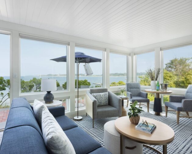 A bright living room with large windows overlooks a body of water at 28 Atlantic in the Wequassett Resort and Golf Club, a renowned Cape Cod wedding venue. Blue sofas, gray chairs, a round coffee table, and indoor plants complete the inviting space.