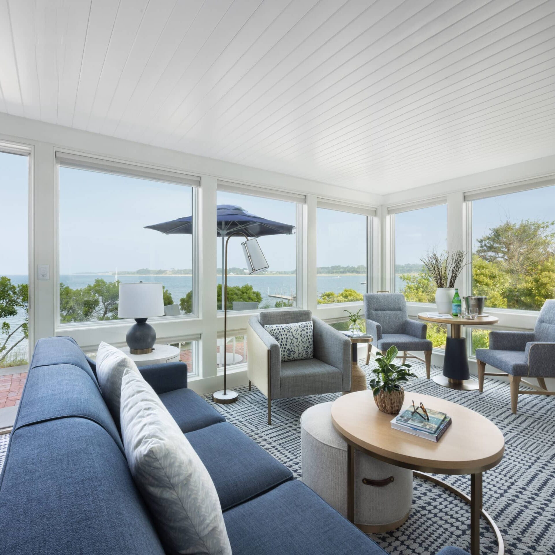 A bright living room with large windows overlooks a body of water at 28 Atlantic in the Wequassett Resort and Golf Club, a renowned Cape Cod wedding venue. Blue sofas, gray chairs, a round coffee table, and indoor plants complete the inviting space.