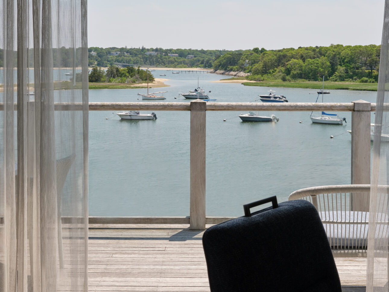 View from a balcony overlooking a calm river with several boats anchored, trees lining the shore, and sheer curtains partially framing this tranquil private coastal escape.