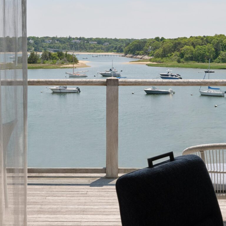 View from a wooden deck overlooking a calm bay with several docked boats, green trees in the background, and part of a chair visible in the foreground—perfect for relaxing after golf at a nearby Cape Cod golf course.